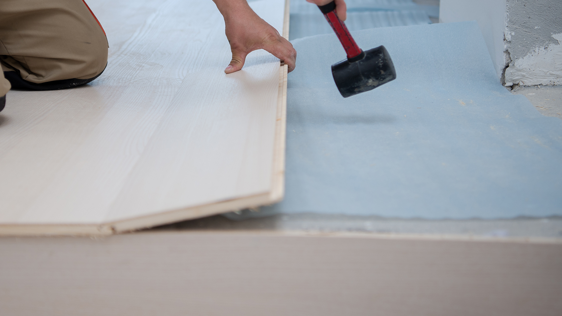 A contractor installing light-colored laminate wood flooring planks using a rubber mallet over a blue foam underlayment.