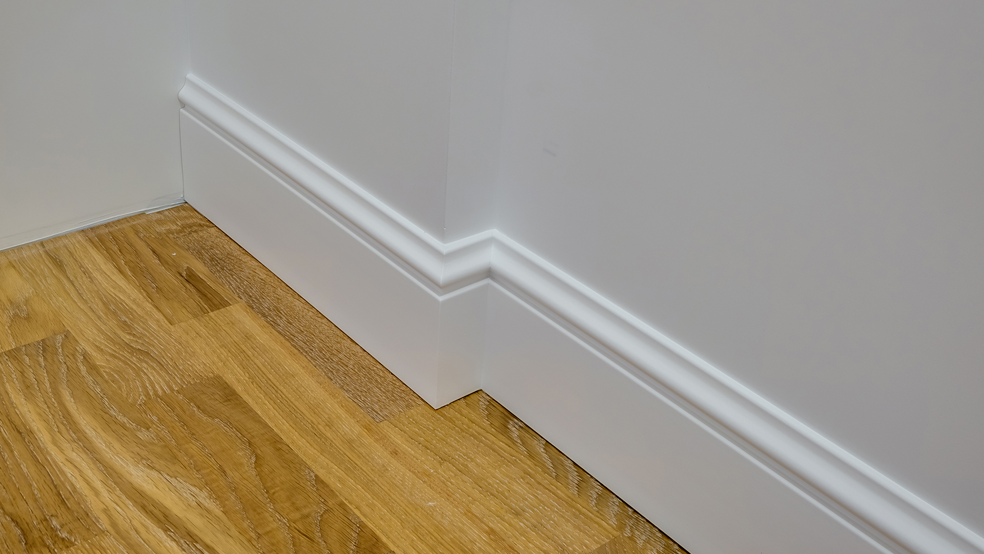Close-up of a professionally installed white decorative baseboard trim on an outside corner with light oak hardwood flooring.